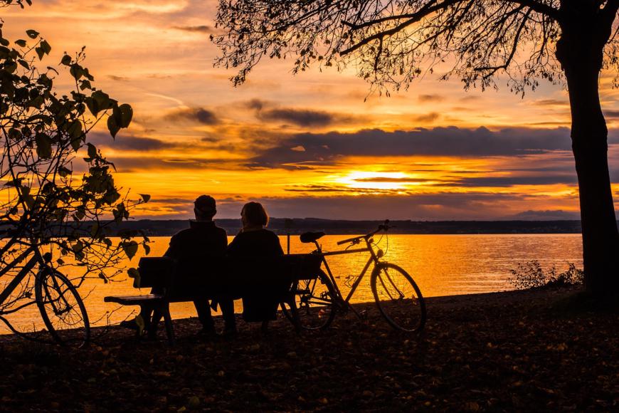 Ein Paar sitzt auf einer Bank und rastet. Fahrräder stehen neben ihnen und sie schauen einen prächtigen Sonnenuntergang an.